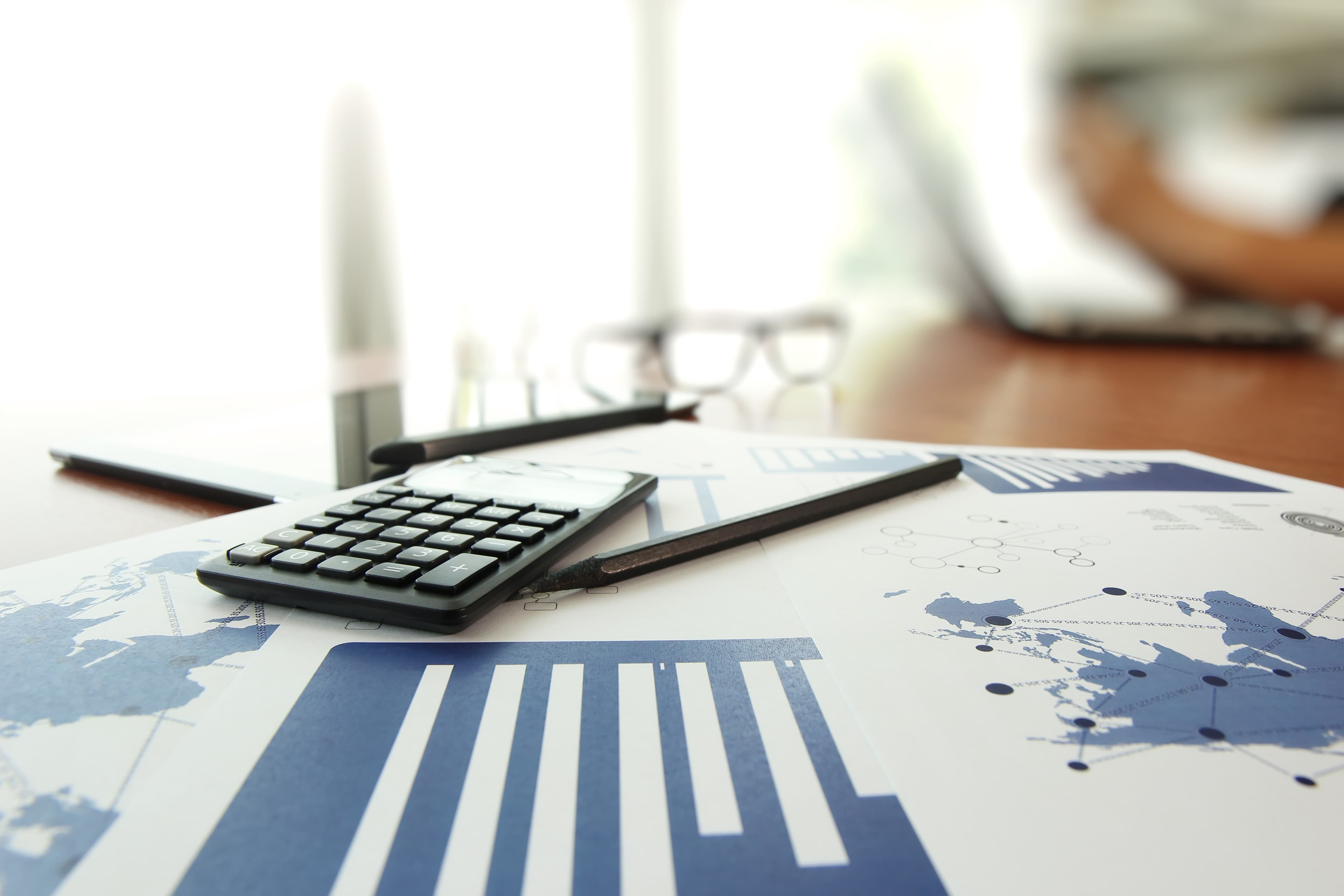 A calculator resting on printed bar charts and world-map data, with glasses and a pen on a wooden desk.